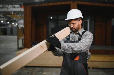 Arap inşaat işçisi ahşap kalas taşıyor, Modüler ev üretim atölyesinde prefabrik ev üzerinde çalışıyor.