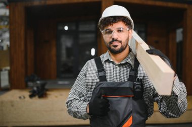 Kendine güvenen Arap marangoz omuzunda ahşap bir kalas taşıyor ve el hareketleriyle işaret ediyor prefabrik bir ev atölyesinde duruyor.