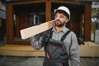 Arap inşaat işçisi omuzunda ahşap kalas taşıyor, prefabrik modüler ev üretim atölyesinde çalışıyor.