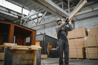 Arap inşaat işçisi prefabrik modüler ev fabrikasında ahşap kalas taşıyor, iş tulumu ve şapka takıyor.