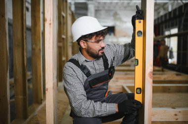 Arap inşaat işçisi, bir atölyede modüler ev inşaatı sırasında ahşap kirişin dikey hizalanmasını sağlamak için ruh seviyesini kullanıyor.