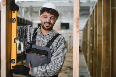 Gülümseyen marangoz, prefabrik ev inşaatı atölyesinde kereste iskeletinin dikey hizalanmasını sağlamak için ruh seviyesini kullanıyor.