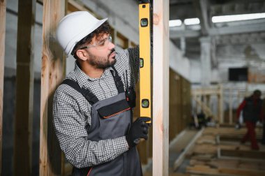 Arap inşaat işçisi prefabrik ev atölyesindeki ruh seviyesini kullanarak ahşap kirişin dikeyliğini kontrol ediyor.