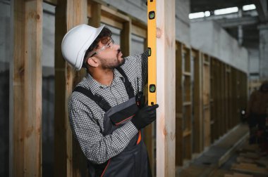 Arap inşaat işçisi, parlak bir atölyede modüler ev yapımı sırasında ahşap kirişin dikey hizalanmasını sağlayan bir ruh seviyesi kullanıyor.