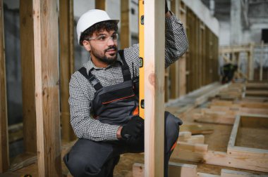 Arap inşaat işçisi inşaat atölyesindeki modüler ev yapımı sırasında ruh seviyesini kullanarak ahşap kirişin dikeyliğini kontrol ediyor.