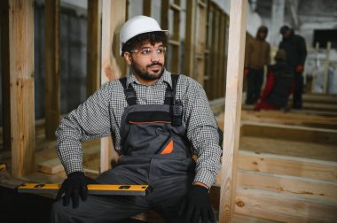 İnşaat işçisi mola veriyor, atölyede ahşap bir çerçevede oturuyor, ruh seviyesini tutuyor, diğer işçiler arka planda meşgulken.