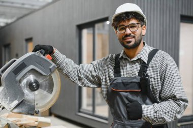 Prefabrik ev atölyesinde dairesel testereyle poz veren kendinden emin inşaat işçisi, modüler binalarda uzmanlık ve güvenlik gösteriyor