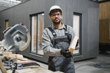 Yetenekli marangoz ahşap kalasları inceliyor, modern fabrikada prefabrik ev projesi üzerinde çalışıyor.