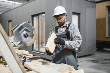 Becerikli marangoz, ahşap kalası dikkatle inceliyor, prefabrik ev inşaatı atölyesini kesmeye hazırlanıyor.