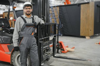 Modüler bir ev üretim tesisinde forklifte yaslanan, tulumlu ve kasklı, kendine güvenen Arap inşaat işçisi.