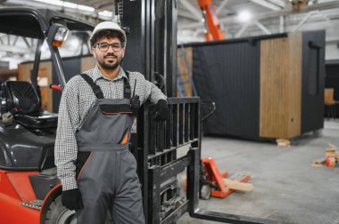 Bir modüler ev üretim fabrikasında forklifte dayanan kalın şapka ve tulum giyen Arap mühendis.