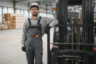 Bir fabrikada ya da büyük bir depoda forklifte yaslanan tulumlu ve kasklı kendine güvenen Arap depo işçisi.