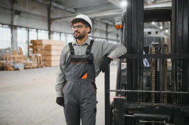 Bir modüler ev yapım atölyesinde forklifte yaslanan güvenlik kaskı ve iş tulumu giyen Arap mühendis.