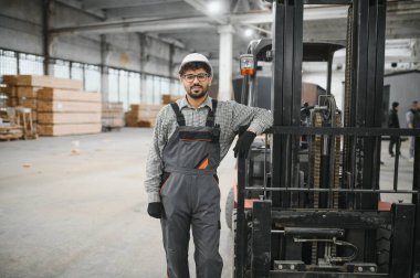 Modüler bir ev üretim tesisinde forklifte yaslanan tulumlu ve kasklı güvenilir Arap depo çalışanı uzmanlık ve profesyonellik gösterisi sergiliyor.