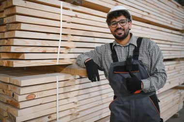 Kendine güvenen Arap marangoz, güvenlik kaskı takıyor. Başparmaklarını kaldırırken marangoz, marangozluk fabrikasındaki kerestelere yaslanıyor ve modüler ev yapımında başarılı oluyor.