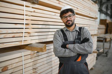 Kendine güvenen Arap marangoz güvenlik kaskı takıyor ve iş tulumları ile kereste deposunda çapraz kollar arasında duruyor, başarılı küçük işletme sahibi.