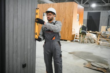 Odaklanmış inşaat işçisi, ruh seviyesini kullanarak, modüler yapı projesinde hassasiyeti güvence altına alarak prefabrik ev panelinin dikey hizasını kontrol ediyor.