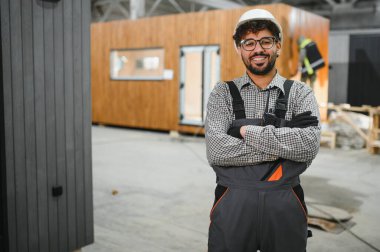 Modüler bir ev yapım atölyesinde çapraz kollarla gülümseyen kendine güvenen Arap inşaat işçisi prefabrik konutlarda uzmanlık ve profesyonellik gösteriyor.