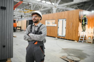 Güvenlik kaskı takan ve iş tulumu giyen Arap inşaat işçisi prefabrik bir ev yapımı atölyesinde çapraz kollarla gülümsüyor.