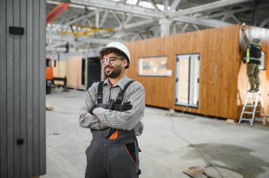 Modüler bir ev fabrikasında çapraz kollarla gülümseyen kendine güvenen Arap inşaat işçisi, üretim sürecini denetliyor.