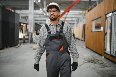 Arap inşaat işçisi bir modüler ev yapım atölyesinde yürüyor, montaj sürecini denetlerken telsizle iletişim kuruyor.