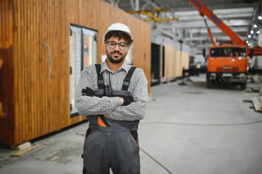 Güvenlik kaskı takan ve iş tulumu giyen kendine güvenen inşaat işçisi modüler ev üretim tesisinde çapraz kollarla ayakta duruyor.