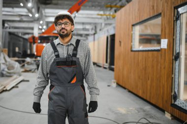 Arap inşaat işçisi, güvenlik kaskı takıyor, elinde telsiz var, büyük bir inşaat atölyesinde prefabrik modüler ev üretimini denetliyor.