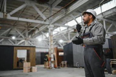 Güvenlik kaskı takan ve telsiz kullanan inşaat işçisi prefabrik ev üretim tesisinde operasyonları koordine ediyor.