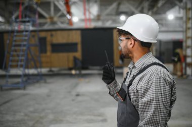 Arap inşaat işçisi telsiz kullanıyor, prefabrik ev imalat tesisinde operasyonları koordine ediyor.