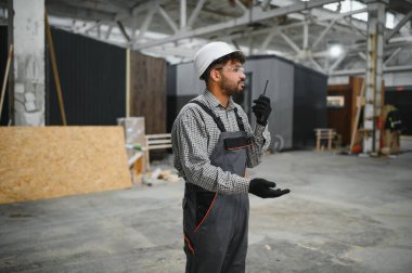 Güvenlik kaskı takan ve telsiz kullanan inşaat işçisi, modüler ev üretim tesisinde operasyonları koordine ediyor.