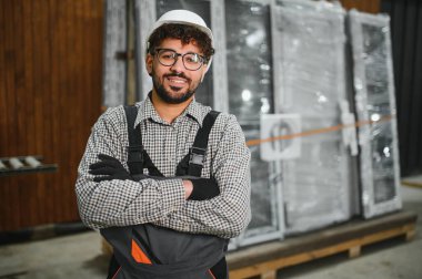 Modüler bir ev yapım atölyesinde çapraz kollarla gülümseyen kendine güvenen Arap inşaat işçisi, prefabrik konut inşaatında uzmanlık ve profesyonellik gösteriyor