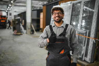 Güvenlik kaskı ve eldiven takan Arap inşaat işçisi modüler bir ev yapım atölyesinde baş parmak işareti yapıyor.