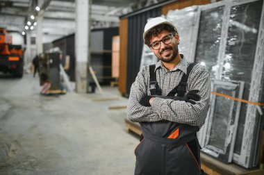 Modüler ev yapım atölyesinde çapraz kollarla gülümseyen kendinden emin bir Arap inşaat işçisinin portresi, prefabrik bina inşaatında uzmanlık ve profesyonellik sergiliyor.