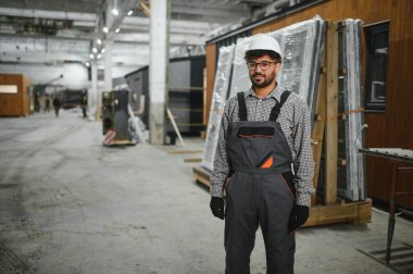 Arap inşaat işçisi, tulum ve şapka giyerek bir atölyede, modüler evlerin üretimini denetliyor.