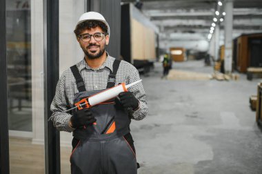 Arap inşaat işçisi, prefabrik ev yapım atölyesinde güvenlik kaskı ve tulum giyerek silikon dolgu tabancası tutuyor.