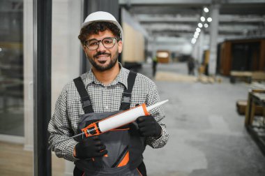 Modüler ev üretim tesisinde çalışan inşaat uzmanı, elinde dolgu malzemesi tutarken gülümsüyor. Arka planda prefabrik elementlerle endüstriyel çalışma alanı