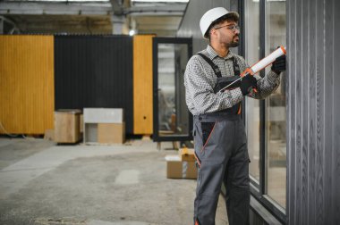 Orta Doğulu inşaat işçisi prefabrik ev fabrikasında pencere çerçevesine dolgu macunu sürerek hava geçirmez ve hava geçirmez tesisat sağlıyor.