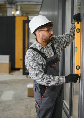 Odaklanmış inşaat işçisi modüler ev duvarının dikey hizalanmasını ruh seviyesini kullanarak, hassas ve kaliteli bir yapı oluşturulmasını sağlayarak kontrol ediyor