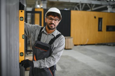 Arap inşaat işçisi, bir modüler ev fabrikasındaki prefabrik duvar bölümünün dikey hizalanmasını sağlıyor.