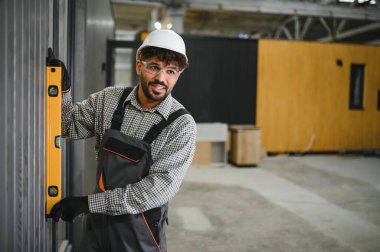 Gülümseyen Arap inşaat işçisi, büyük atölyedeki ruh seviyesini kullanarak prefabrik modüler ev duvarının dikey hizasını kontrol ediyor