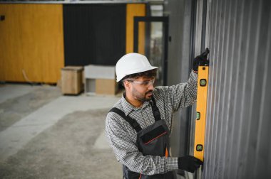 Prefabrikasyon atölyesinde modüler ev montajı sırasında duvar panelinin dikey hizalanmasını sağlamak için ruh seviyesini kullanarak odaklanmış inşaat işçisi