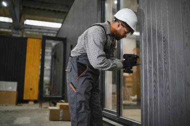 Odaklanmış inşaat işçisi, büyük atölyeye prefabrik modüler ev üzerine duvar paneli kurmak için elektrikli matkap kullanıyor