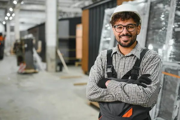 Modüler bir ev yapım atölyesinde çapraz kollarla gülümseyen kendinden emin bir Arap inşaat işçisinin portresi, prefabrik konut inşaatında uzmanlık ve profesyonellik sergiliyor
