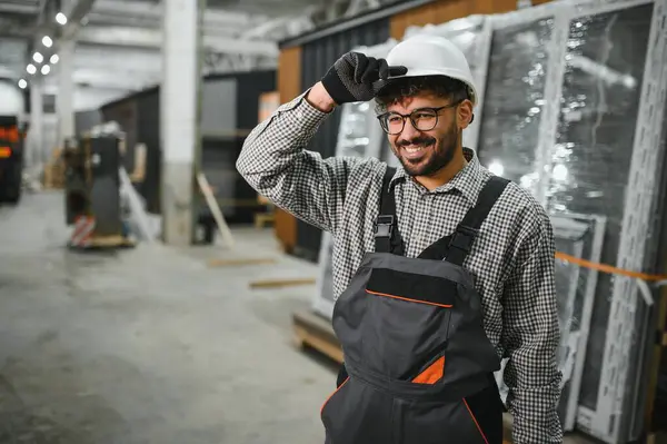 Arap inşaat işçisi, modüler ev yapım atölyesinde şapkasını düzeltirken gülümsüyor, işyerinden memnuniyet ve güvenlik gösterisi sergiliyor