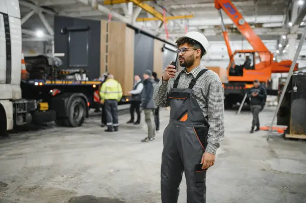 Güvenlik kaskı takan ve telsiz kullanan Arap mühendis modüler bir ev inşaatı atölyesinde operasyonları koordine ediyor.