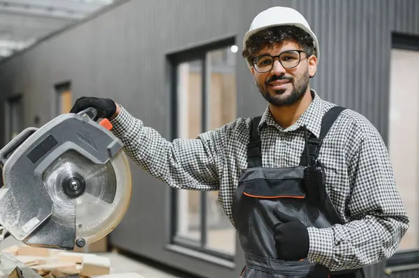 Prefabrik ev atölyesinde dairesel testereyle poz veren kendinden emin inşaat işçisi, modüler binalarda uzmanlık ve güvenlik gösteriyor