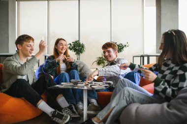 Dört neşeli genç yetişkin rahat armut koltuklarda oturup modern bir oturma odasında masa oyunu oynuyorlar.