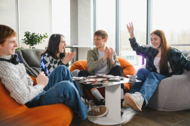 Bir grup neşeli genç arkadaş masa oyunu oynuyor, el çırpıyorlar ve puf minderlerde otururken birlikte eğleniyorlar.