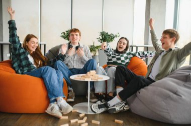 Dört neşeli arkadaş puf minderlerinde oturmuş, Jenga oynuyorlar, kule çökerken heyecan ve mutluluğu ifade ediyorlar.