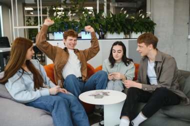 Dört neşeli meslektaşı modern bir çalışma ortamında puf minderlerinde oturuyor ve molalarında kart oyununun keyfini çıkarıyorlar.